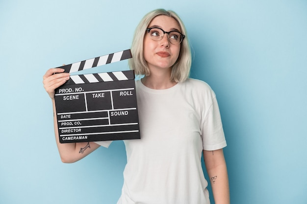 Jonge kaukasische vrouw die een filmklapper houdt die op blauwe achtergrond wordt geïsoleerd en droomt van het bereiken van doelen en doeleinden