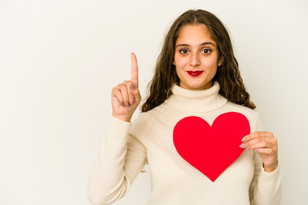 Jonge kaukasische vrouw die een geïsoleerde de dagvorm van hartvalentijnskaarten houdt die nummer één met vinger toont.