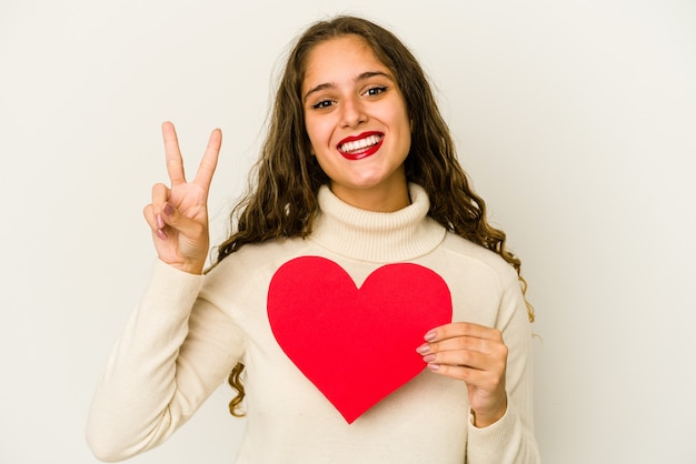 Jonge kaukasische vrouw die een geïsoleerde de dagvorm van hartvalentijnskaarten houdt die nummer twee met vingers toont.