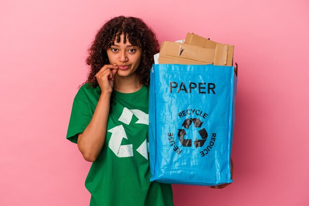 Jonge kaukasische vrouw die een gerecycled plastic houdt dat op roze muur wordt geïsoleerd met vingers op lippen die een geheim houden.