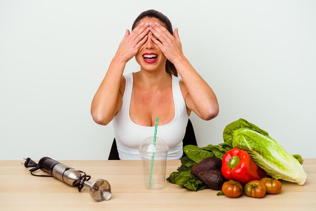 Jonge kaukasische vrouw die een gezonde smoothie met groenten voorbereidt behandelt ogen met handen, glimlacht in het algemeen wachtend op een verrassing.