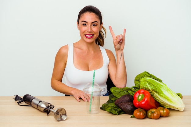 Jonge kaukasische vrouw die een gezonde smoothie met groenten voorbereidt die een hoornengebaar als revolutieconcept tonen.