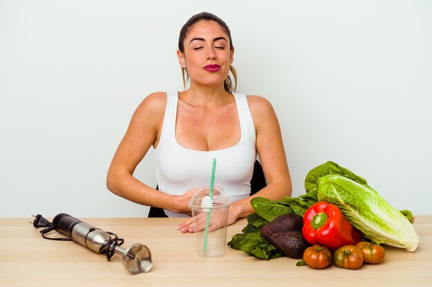 Jonge kaukasische vrouw die een gezonde smoothie met groenten voorbereidt raakt buik, glimlacht zachtjes, eet en tevredenheidsconcept.