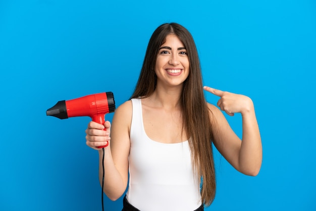 Jonge kaukasische vrouw die een haardroger houdt die op blauwe achtergrond wordt geïsoleerd en een duim omhoog gebaar geeft