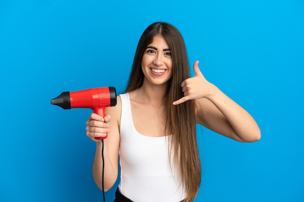 Jonge kaukasische vrouw die een haardroger houdt die op blauwe muur wordt geïsoleerd en telefoongebaar maakt