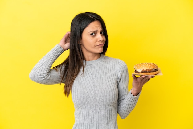 Jonge kaukasische vrouw die een hamburger houdt die op gele achtergrond wordt geïsoleerd die twijfels heeft