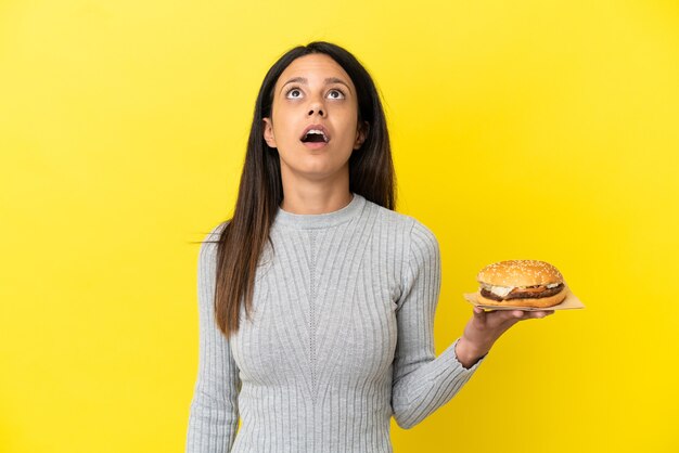 Jonge kaukasische vrouw die een hamburger houdt die op gele achtergrond wordt geïsoleerd en omhoog kijkt en met verbaasde uitdrukking