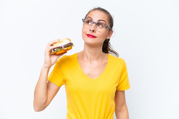 Jonge kaukasische vrouw die een hamburger houdt die op witte achtergrond wordt geïsoleerd en omhoog kijkt