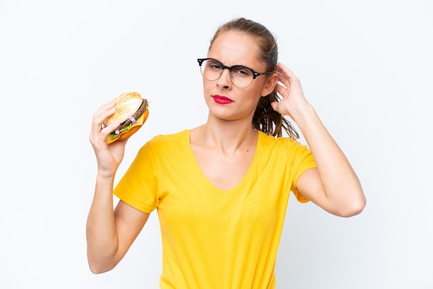 Jonge kaukasische vrouw die een hamburger houdt die op witte achtergrond wordt geïsoleerd en twijfels heeft