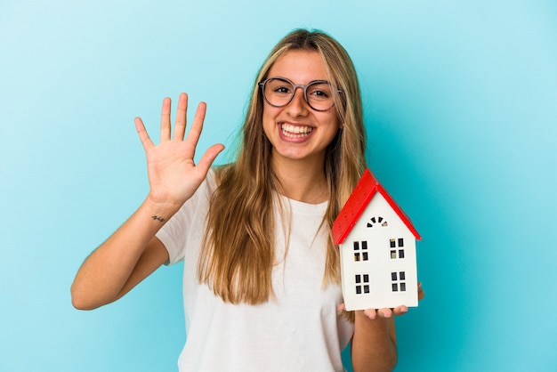 Jonge kaukasische vrouw die een huismodel houdt dat op blauwe muur vrolijk glimlachen toont die nummer vijf met vingers toont