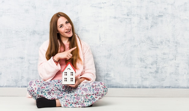 Jonge Kaukasische vrouw die een huispictogram houdt glimlachend vrolijk wijzend met weg wijsvinger.