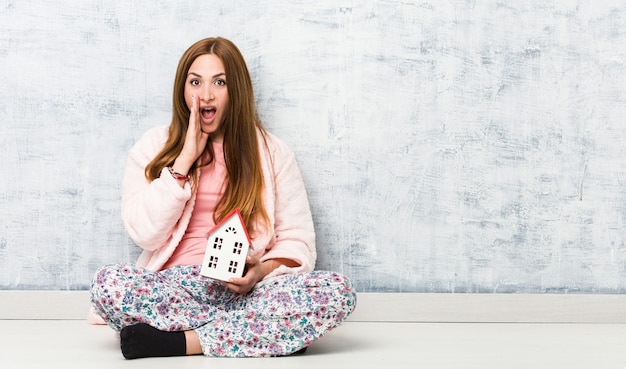 Jonge kaukasische vrouw die een huispictogram houdt opgewonden aan voorzijde schreeuwt.