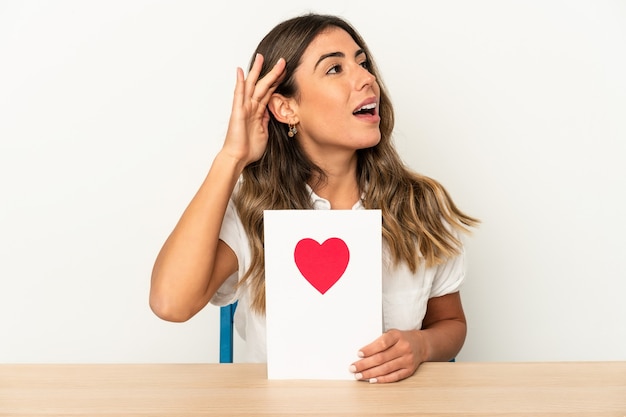 Jonge kaukasische vrouw die een kaart van de valentijnskaartendag houdt die geïsoleerd probeert te luisteren een roddel.