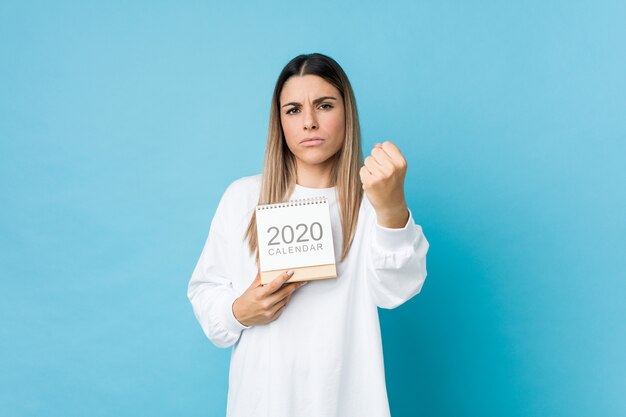 Jonge kaukasische vrouw die een kalender van 2020 houdt die vuist toont aan met agressieve gelaatsuitdrukking.