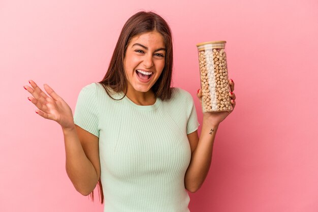 Jonge kaukasische vrouw die een kikkererwtenkruik houdt die op roze achtergrond wordt geïsoleerd en een aangename verrassing ontvangt, opgewonden en handen opsteekt.