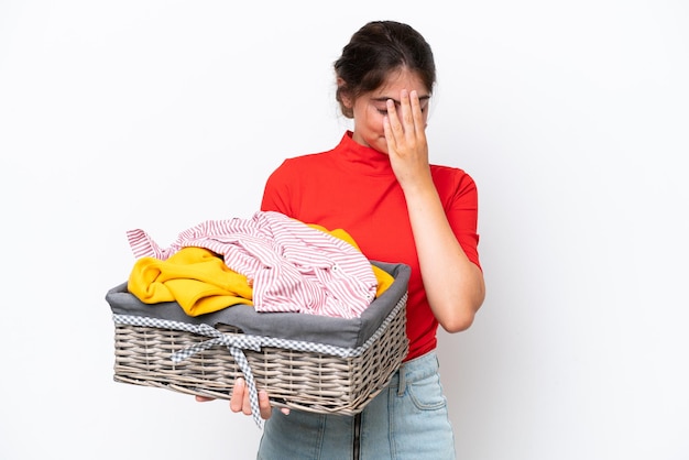 Jonge kaukasische vrouw die een klerenmand houdt die op witte achtergrond met vermoeide en zieke uitdrukking wordt geïsoleerd