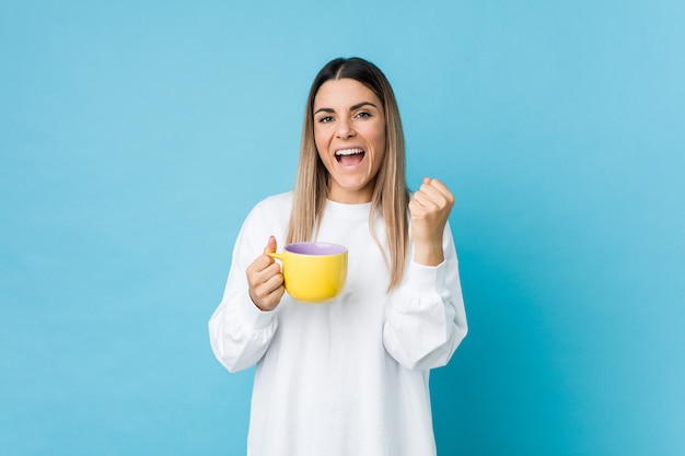 Jonge Kaukasische vrouw die een koffiekop houden die onbezorgd en opgewekt toejuichen. Overwinning concept.