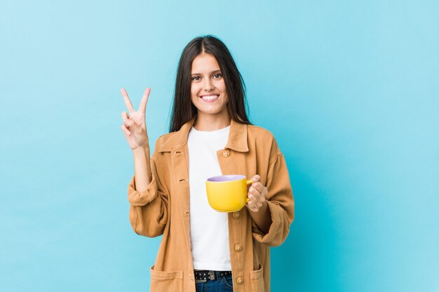 Jonge Kaukasische vrouw die een koffiemok houdt die nummer twee met vingers toont.