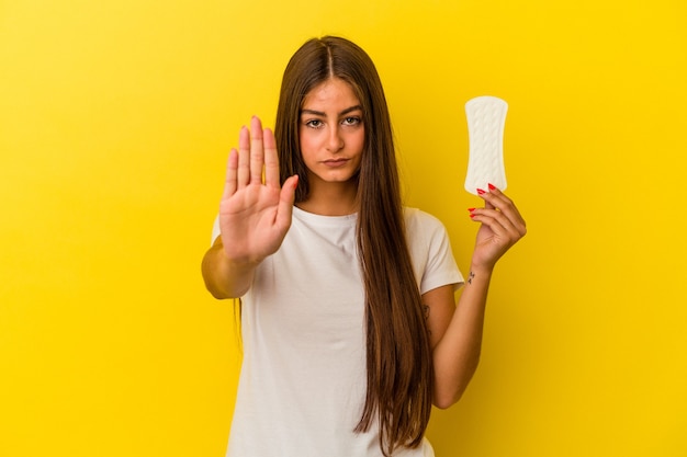 Jonge kaukasische vrouw die een kompres houdt dat op gele achtergrond wordt geïsoleerdd die zich met uitgestrekte hand bevindt die stopteken toont, die u verhinderen.