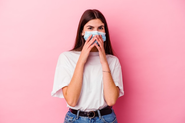 Jonge kaukasische vrouw die een masker voor virus draagt dat op roze achtergrond wordt geïsoleerd die over iets lacht, mond bedekt met handen.