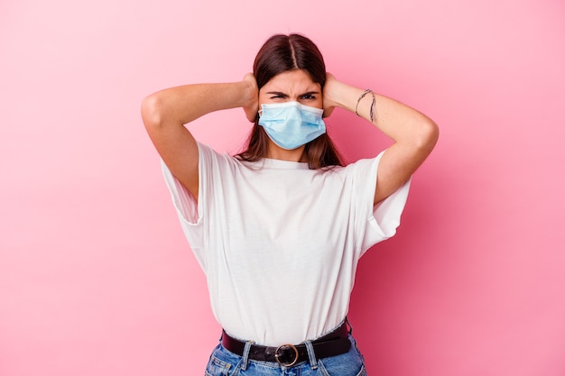 Jonge kaukasische vrouw die een masker voor virus op roze draagt die oren bedekt met handen die niet te hard geluid proberen te horen.