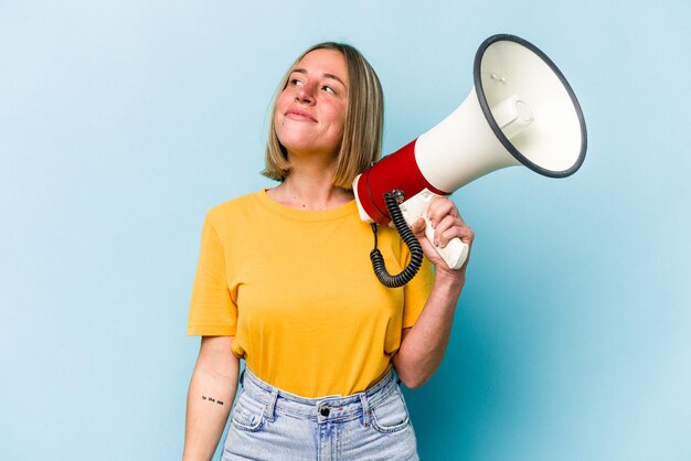 Jonge kaukasische vrouw die een megafoon houdt die op blauwe achtergrond wordt geïsoleerd en droomt van het bereiken van doelen en doeleinden