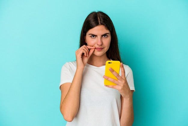 Jonge kaukasische vrouw die een mobiele telefoon houdt die op blauwe achtergrond met vingers op lippen wordt geïsoleerd die een geheim houden.