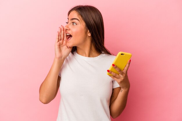 Jonge kaukasische vrouw die een mobiele telefoon houdt die op roze achtergrond wordt geïsoleerd die schreeuwt en palm dichtbij geopende mond houdt.