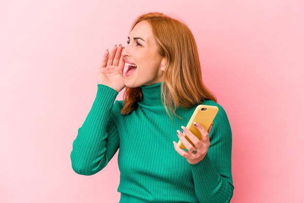 Jonge kaukasische vrouw die een mobiele telefoon houdt die op roze achtergrond wordt geïsoleerd die schreeuwt en palm dichtbij geopende mond houdt.