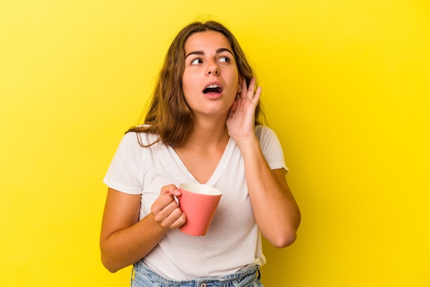 Jonge kaukasische vrouw die een mok houdt die op gele achtergrond wordt geïsoleerd die probeert om een roddel te luisteren.