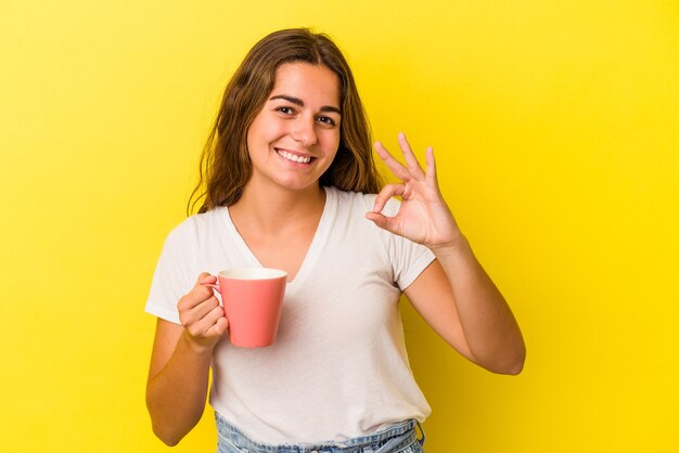 Jonge kaukasische vrouw die een mok houdt die op gele achtergrond wordt geïsoleerd, vrolijk en zelfverzekerd met een goed gebaar.