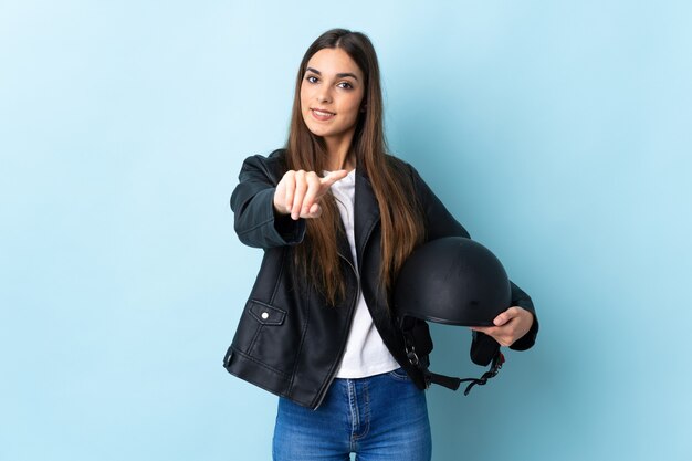 Jonge kaukasische vrouw die een motorhelm houdt die op blauw richtend front met gelukkige uitdrukking wordt geïsoleerd