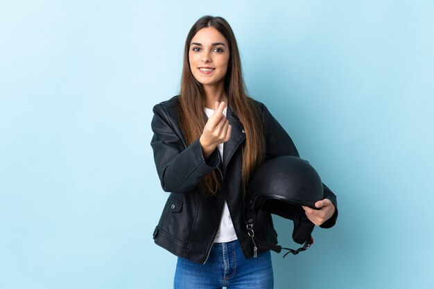 Jonge kaukasische vrouw die een motorhelm houdt die op blauwe muur wordt geïsoleerd die geldgebaar maakt