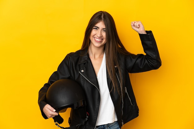 Jonge kaukasische vrouw die een motorhelm houdt die op gele achtergrond wordt geïsoleerd en sterk gebaar doet