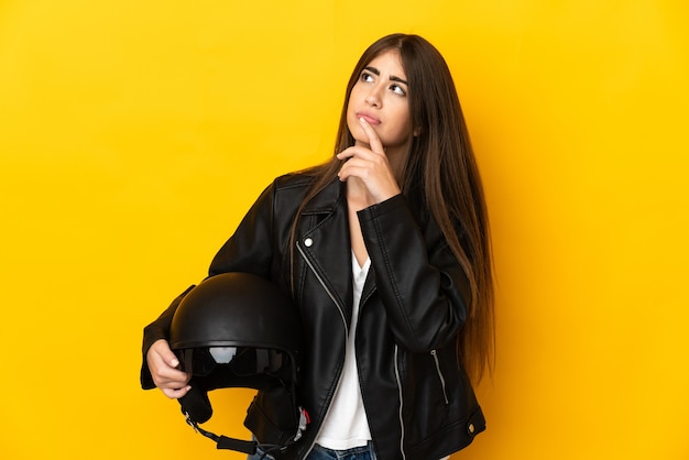 Jonge kaukasische vrouw die een motorhelm houdt die op gele muur wordt geïsoleerd die twijfels heeft tijdens het kijken