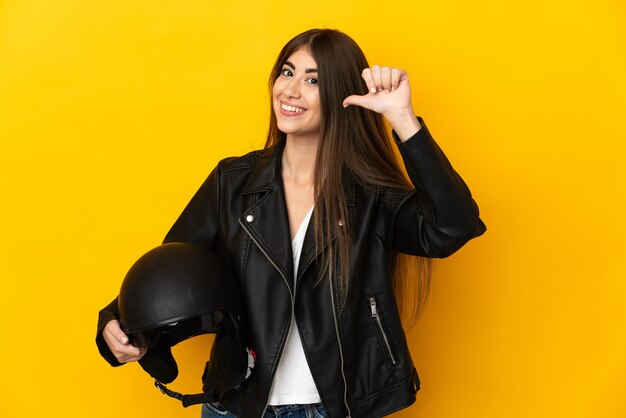 Jonge kaukasische vrouw die een motorhelm houdt die op gele muur wordt geïsoleerd, trots en zelfvoldaan