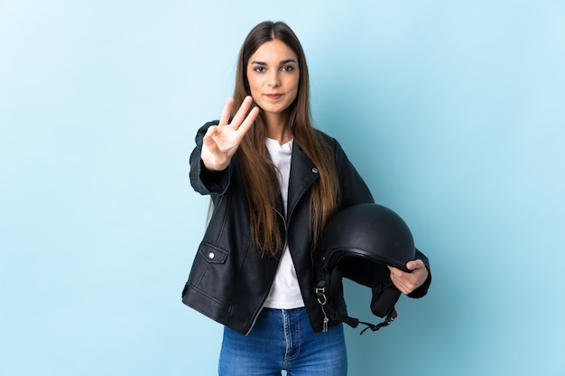 Jonge kaukasische vrouw die een motorhelm op gelukkig blauw houdt en drie met vingers telt