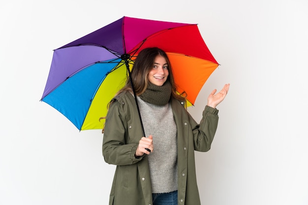 Jonge kaukasische vrouw die een paraplu houdt die op witte muur wordt geïsoleerd die handen uitstrekt naar de zijkant om uit te nodigen om te komen