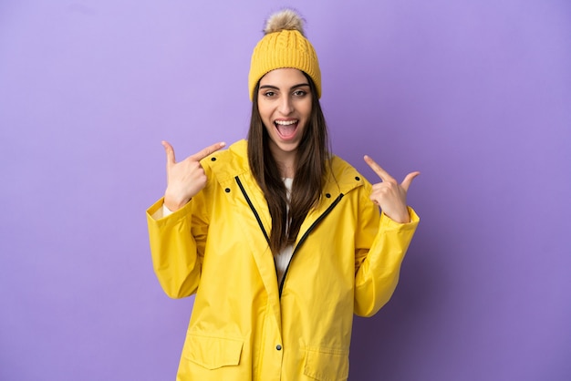 Jonge kaukasische vrouw die een regendichte jas draagt die op paarse achtergrond wordt geïsoleerd en een duim omhoog gebaar geeft