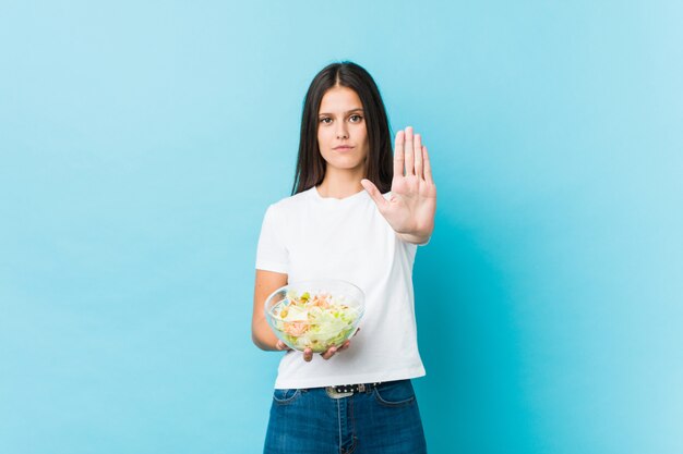 Jonge Kaukasische vrouw die een salade houdt die zich met uitgestrekte hand bevindt die eindeteken toont, dat u verhindert.