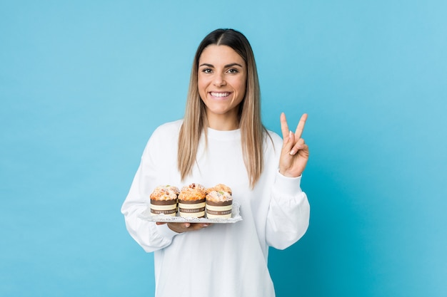 Jonge Kaukasische vrouw die een snoepjescake houdt die overwinningsteken toont en breed glimlacht.