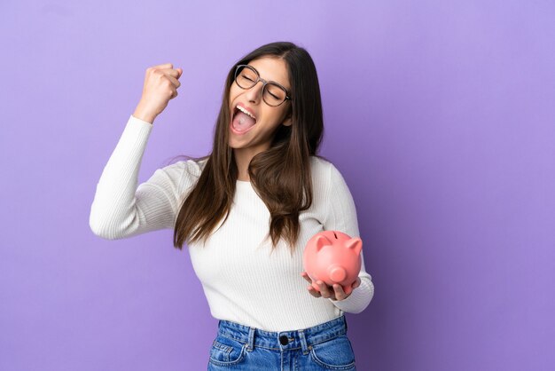 Jonge kaukasische vrouw die een spaarvarken houdt dat op purpere achtergrond wordt geïsoleerd en een overwinning viert