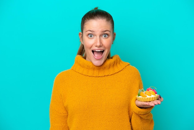 Jonge kaukasische vrouw die een taartje houdt dat op blauwe achtergrond met verrassing en geschokte gezichtsuitdrukking wordt geïsoleerd