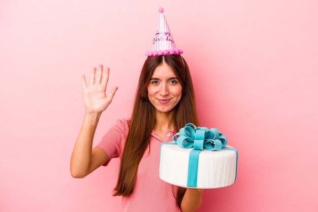Jonge kaukasische vrouw die een verjaardag viert die op roze achtergrond wordt geïsoleerd die vrolijk glimlacht die nummer vijf met vingers toont.