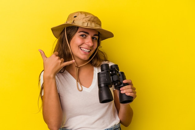 Jonge kaukasische vrouw die een verrekijker houdt die op gele achtergrond wordt geïsoleerd die een mobiel telefoongesprekgebaar met vingers toont.