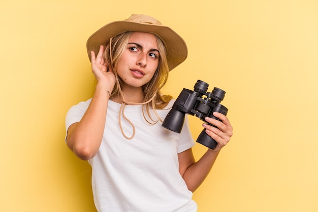 Jonge kaukasische vrouw die een verrekijker houdt die op gele achtergrond wordt geïsoleerd die probeert om een roddel te luisteren.