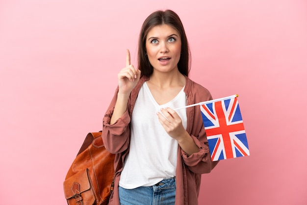 Jonge kaukasische vrouw die een vlag van het Verenigd Koninkrijk houdt die op roze achtergrond wordt geïsoleerd en een idee denkt dat de vinger omhoog wijst