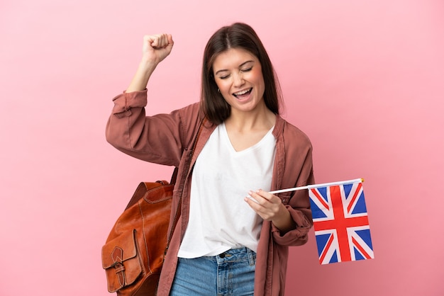 Jonge kaukasische vrouw die een vlag van het Verenigd Koninkrijk houdt die op roze achtergrond wordt geïsoleerd en een overwinning viert