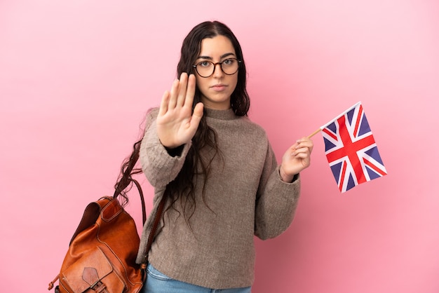 Jonge kaukasische vrouw die een vlag van het Verenigd Koninkrijk houdt die op roze achtergrond wordt geïsoleerd en stopgebaar maakt