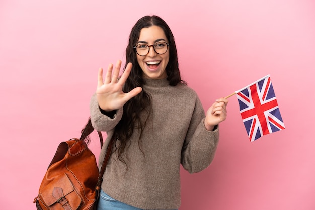 Jonge kaukasische vrouw die een vlag van het Verenigd Koninkrijk houdt die op roze achtergrond wordt geïsoleerd en vijf met vingers telt